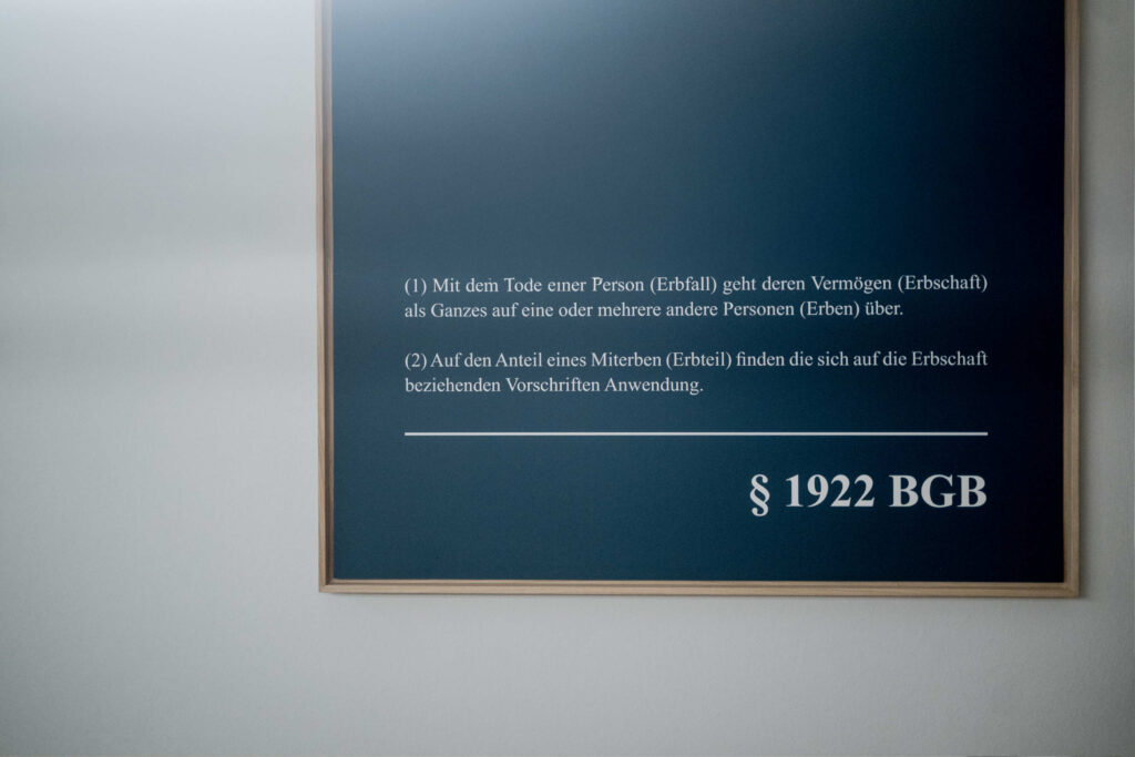 Rechteckige Tafel mit dunkelblauer Oberfläche und hellem Rahmen. Weiße Schrift in serifenloser Typografie zeigt einen Gesetzestext mit zwei nummerierten Absätzen und einer Paragraphenangabe. Die Tafel hängt an einer hellen Wand.